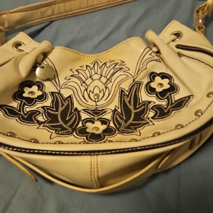 Beige axelväska med blommigt mönster - Säljer en beige axelväska i skinn med svart broderat blommönster och nitar framtill. Väskan har justerbar axelrem med metallspännen och dekorativa detaljer. Perfekt för dig som gillar unika accessoarer med lite extra attityd.