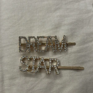 Glittriga hårspännen med text - Två snygga hårspännen i guld med glittrande stenar som bildar orden DREAM och STAR. Perfekta för att ge frisyren extra bling och attityd. Bokstäverna är täckta av gnistrande stenar som verkligen fångar ljuset.