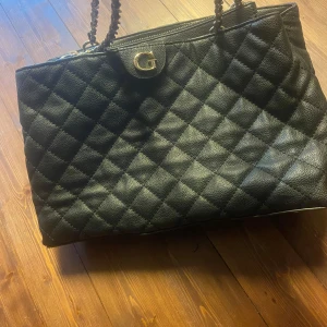 Svart quiltad handväska från Guess - Säljer en svart quiltad handväska från Guess med kedjehandtag och guldfärgade detaljer. Väskan är tillverkad i skinnimitation och har dragkedja baktill samt ett stort G i metall framtill. Insidan är ljus med Guess-logga. Perfekt för dig som gillar stilrena accessoarer.