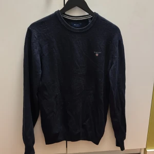 Mörkblå tröja från Gant - Klassisk mörkblå tröja från Gant med rund halsringning och diskret broderad logga på bröstet. Tröjan har långa ärmar och ribbade muddar vid ärmslut och nederkant. Perfekt för en stilren och enkel look.