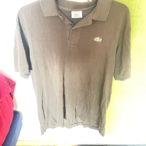 Brun pikétröja från Lacoste - Brun pikétröja från Lacoste med klassisk krage och korta ärmar. Tröjan har den ikoniska gröna krokodilloggan broderad på bröstet och är tillverkad i bomull. Perfekt för en avslappnad och stilren look.