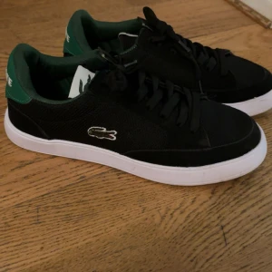 Svarta lacoste skor  - Snygga svarta Lacoste sneakers med gröna detaljer på häl och insida. Skorna har vit sula, snörning och klassisk krokodillogga på sidan. Ovandelen är i mesh och mocka för en sportig look. Perfekta för dig som gillar stilrena och sportiga skor.