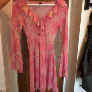 Rosa blommig omlottklänning med volang - Supersöt rosa kortklänning med blommigt mönster och volangkant längs v-ringningen. Klänningen har långa utsvängda ärmar och knytband framtill. Tunt, lätt material som ger en flowy känsla. Perfekt för vår och sommar!