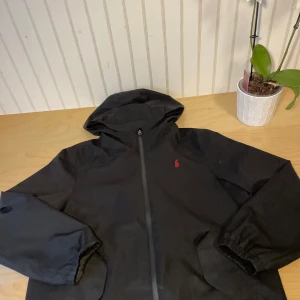 Svart Polo Ralph Lauren Regnjacka - Svart vindjacka från Polo Ralph Lauren med huva och dragkedja framtill. Jackan har röd broderad logga på bröstet, två fickor med knappstängning och elastiska muddar. Tillverkad i polyester och perfekt för blåsiga dagar.