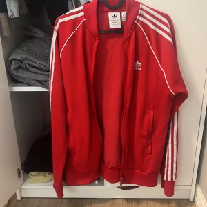 Röd Adidas track jacket med vita ränder - Riktigt snygg röd Adidas track jacket med klassiska vita ränder längs ärmarna och vit logga på bröstet. Jackan har dragkedja framtill, ribbade muddar och är tillverkad i återvunnen polyester (Primeblue). Perfekt för en sportig och chill stil.