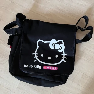 Hello Kitty svart axelväska - Svart axelväska med stort Hello Kitty-tryck på framsidan och justerbar axelrem. Väskan har ett stort huvudfack med dragkedja och extra innerficka. Materialet är slitstark polyester och designen är enkel men söt med vit och rosa detalj.