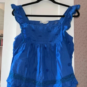 Blå blus med volang från MNG - Supersöt blå blus från MNG med broderade blommor och volangdetaljer vid axelbanden och nederkanten. Blusen är ärmlös och har en luftig passform, perfekt för varma dagar. Tillverkad i bomull och har fina hålbroderier som ger en extra touch.