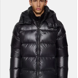 Svart pufferjacka från J.Lindeberg - Snygg svart pufferjacka från J.Lindeberg med hög krage, huva och dragkedja framtill. Jackan har en glansig finish, breda kanaler och diskret logga på bröstet. Perfekt för kalla dagar när du vill hålla stilen.