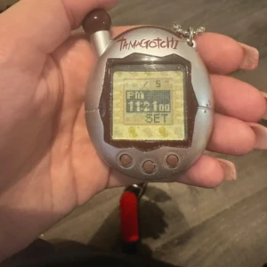 Tamagotchi V4 - Klassisk Tamagotchi digitalt husdjur från Bandai. Silverfärgad modell med tre knappar och tydlig display. Något slitage syns på ytan men fungerar som den ska. Perfekt för samlare eller nostalgiker som vill återuppleva 90-talets digitala husdjursvåg.