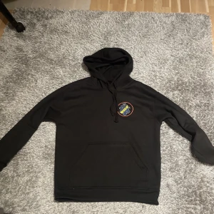 Aik hoodie (HATA ALLA) - Svart hoodie med huva och snörning, stor magficka och tryck på bröstet samt stort gult tryck 'HATA ALLA' på ryggen. Perfekt för dig som vill sticka ut. Mjuk och skön, i bomull. Hör av er vid frågor & funderingar 