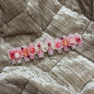 Rosa dekorerade lösnaglar med glitter - Säljer ett set lösnaglar i rosa och ljusrosa med glitter, pärlor, 3D-blommor, rosetter och strass. Naglarna har en kistform och är dekorerade med både små och stora detaljer i vitt, rosa och orange. Perfekt för dig som vill ha extra glam på naglarna.