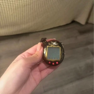 Tamagotchi V5 - Klassisk Tamagotchi digitalt husdjur från Bandai. Svart med gulddetaljer och tre röda knappar. Skärmen och knapparna är hela ut, små tecken på användning men inga skador. Fungerar perfekt. Perfekt för samlare eller nostalgiker som vill återuppleva 90-talet.