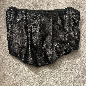 Svart paljettkorsett med snörning - Säljer en svart glittrig korsett-topp täckt av paljetter. Toppen har snörning i ryggen för justerbar passform och en snygg, croppad look. Perfekt för dig som vill sticka ut med en festlig vibe. Materialet är syntet och toppen är axelbandslös.