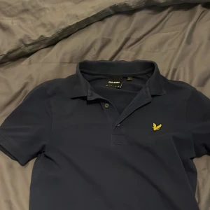 Marinblå Lyle & Scott pikétröja - Snygg marinblå pikétröja från Lyle & Scott med klassisk krage och korta ärmar. Tröjan har gul broderad logga på bröstet och är gjord i mjuk bomull. Perfekt för dig som gillar stilrena och tidlösa plagg.