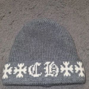 Grå Chrome Hearts mössa - Grå ribbstickad mössa med uppvikt kant. På framsidan finns vita broderade kors och bokstäver i gotisk stil. Mjuk och stretchig, perfekt för kyliga dagar. Passar dig som gillar streetwear och vill ha en stilren men ändå unik accessoar.