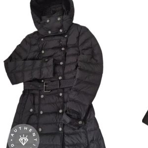 Svart dunjacka från Burberry Brit women xs - Snygg svart dunjacka från Burberry Brit med avtagbar huva, dubbelknäppning och bälte i midjan. Jackan har quiltad design, långa ärmar och klassiskt rutigt foder. Perfekt för kalla dagar när du vill vara både varm och stilren.