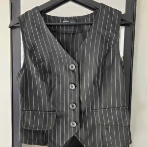 Randig väst från SHEIN med knappar - Snygg svart väst från SHEIN med vita vertikala ränder och fyra knappar framtill. Västen har två fickor och v-ringad hals. Perfekt för att ge outfiten en stilren touch.