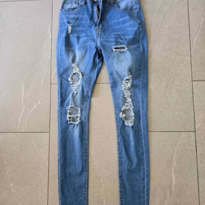 Blå skinny jeans med slitningar - Snygga blå skinny jeans med flera slitningar och trasiga detaljer på benen. Jeansen har klassisk femficksdesign och är tillverkade i stretchigt denim för en tight passform. Perfekta för en trendig och avslappnad stil.