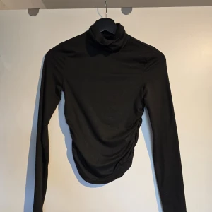 Svart polotopp med rynkade detaljer - Säljer en svart polotopp med långa ärmar och snygga rynkade detaljer på ena sidan. Toppen har en tight passform och är tillverkad i ett stretchigt, mjukt material som sitter skönt på kroppen. Perfekt för dig som gillar stilrena och trendiga plagg.