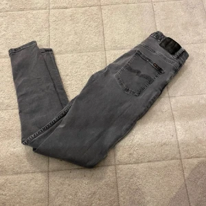 Grå jeans från Nudie Jeans - Säljer ett par grå jeans från Nudie Jeans med klassisk femficksdesign och svarta detaljer på bakfickan. Jeansen har en rak passform och är tillverkade i mjukt denimtyg som sitter skönt hela dagen.
