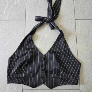 Svart randig omlottväst med knyt - Trendig svart väst med vita ränder och knyt i nacken. Omlottmodell med tre knappar framtill och två dekorativa fickor. Perfekt för att lyfta en outfit med en edgy touch.