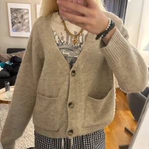 Beige kofta från AWARE med fickor - Supermysig beige kofta från AWARE med v-ringning, stora knappar och två rejäla fickor framtill. Koftan är stickad och har en loose passform med långa ärmar och ribbade muddar. Perfekt att slänga över en t-shirt för en chill look.