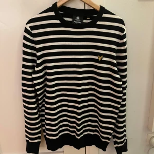 Randig tröja från Lyle & Scott - Snygg svartvit randig tröja från Lyle & Scott med klassisk rund hals och broderad gul logga på bröstet. Tröjan är långärmad och har ribbade muddar vid ärmslut och nederkant. Perfekt för dig som gillar stilren och tidlös design.