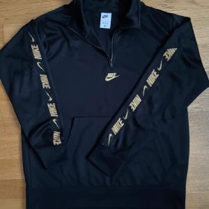 Svart Nike half zip hoodie L - Svart hoodie från Nike med guldiga loggor längs ärmarna och på bröstet. Tröjan har half zip, huva och känguruficka. Tillverkad i mjuk polyester, perfekt för en sportig och chill stil.