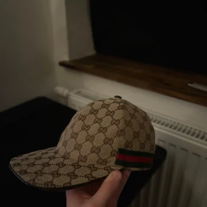 Gucci keps med GG-mönster  - Snygg keps från Gucci i beige med klassiskt GG-monogram över hela kepsen. Kepsen har en böjd skärm och en ikonisk röd och grön rand på sidan. Tillverkad i canvasliknande material med justerbart spänne bak och svart insida med Gucci-logga. Vid snabbaffär kan priset diskuteras! 