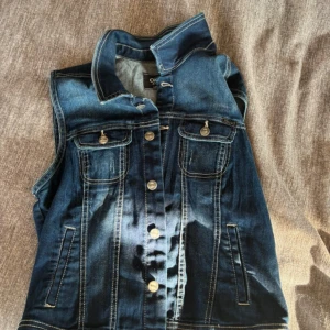 Jeansväst från ONLY i blå denim - Snygg jeansväst från ONLY i klassisk blå denim med kontrastsömmar och knappar framtill. Västen har två bröstfickor med lock och knapp samt två sidofickor. Perfekt att slänga över en hoodie eller t-shirt. Den är i storlek L, men sitter perfekt på mig som vanligtvis är en S/M. Frakt ingår ej💕
