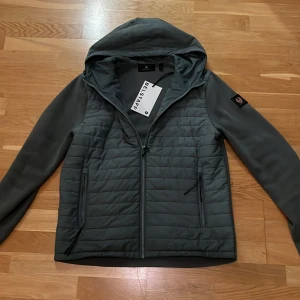 Cardigan  - Snygg mineralgrön cardigan  från Belstaff med quiltad front, huva och dragkedja. Jackan har släta ärmar, två fickor med dragkedja och en patch på ena ärmen. Perfekt för dig som vill ha en stilren och sportig look. Nypris: 4099 kr 
