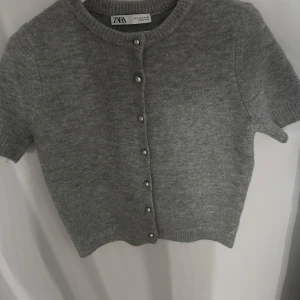 Grå kofta från Zara med pärlknappar - Kortärmad grå kofta från Zara i mjukt stickat material. Snygga pärlknappar framtill ger en extra touch. Perfekt att slänga över en topp eller klänning för en chill look.