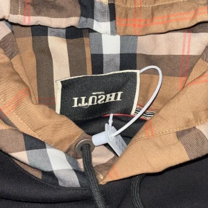 Svart hoodie med burberry hoodie - Svart hoodie från ILQSHI med en snygg rutig huva i beige, brunt och svart. Huva med snörning och metalländar, där ena änden har en B-detalj. Stor magficka framtill och klassisk passform. Perfekt för dig som gillar streetstyle och vill sticka ut.