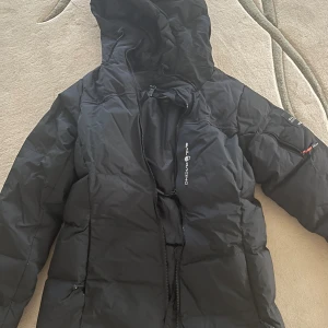 Svart Sail Racing dunjacka med huva + en puma jacka på köpet - Säljer en svart dunjacka från Sail Racing, modell Antarctica Expedition. Jackan har huva, dragkedja framtill och snygga logodetaljer på bröstet och ärmen. Perfekt för kalla dagar och har en cool expedition-vibe. Fickor med dragkedja och stilren design. Använts ändast 2-3 gånger