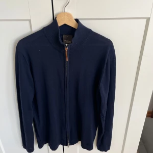 Mörkblå kofta från Oscar Jacobson - Mörkblå kofta från Oscar Jacobson med hel dragkedja och hög krage. Tillverkad i 100% merino ull med lång ärm och stilren look. Perfekt för lager-på-lager och passar till jeans. 