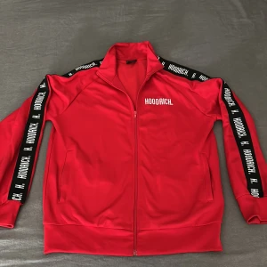 Röd Hoodrich ziptröja med logga - Röd ziptröja från Hoodrich med vit logga på bröstet och svarta band med vit text längs ärmarna. Tröjan har hög krage, hel dragkedja och två fickor framtill. Tillverkad i mjukt och sportigt polyestermaterial, perfekt för en streetwear-look. Tröjan har endast används 5 gånger ungefär