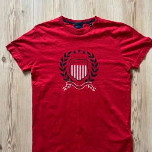Röd GANT t-shirt med emblem, stl S - Röd t-shirt från GANT med klassiskt emblemtryck framtill i svart och vitt. T-shirten har rund halsringning och korta ärmar. Tillverkad i mjuk bomull för en skön känsla. Perfekt för dig som gillar preppy och stilren design.