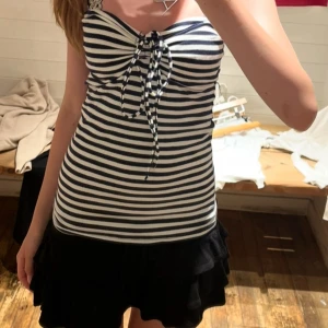 Randig bandeau topp med knyt - Säljer en snygg bandeau topp med marinblå och vita ränder. Toppen har knyt framtill och är ärmlös, perfekt för sommarens varma dagar. Den är figurnära och har en klassisk, fräsch look som passar till både kjol och jeans.