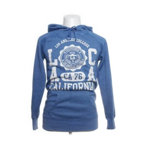 Blå hoodie Los Angeles College - Blå hoodie från Los Angeles College med vit tryckt text och emblem framtill. Tröjan har huva med snörning och en stor magficka. Perfekt för en chill och avslappnad stil. Långärmad och i mjukt material.