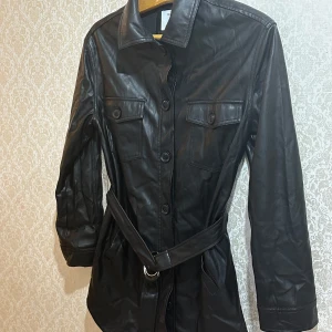 Svart skinnjacka med bälte från Bershka - Snygg svart skinnjacka från Bershka med bälte i midjan och knappar framtill. Jackan har två bröstfickor med lock och långa ärmar. Perfekt för dig som vill ha en edgy och trendig look.