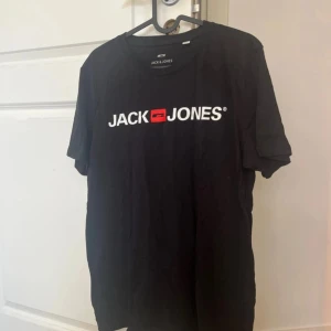 Svart Jack & Jones t-shirt i bomull - Svart t-shirt från Jack & Jones med vit och röd logotyp tryckt på bröstet. Klassisk rund hals och korta ärmar. Tillverkad i mjuk bomull som är skön mot huden. Perfekt för dig som gillar enkel och stilren streetwear.