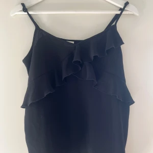 Svart volanglinne från Jacqueline de Yong - Snygg svart linnetopp från Jacqueline de Yong med tunna axelband och volangdetaljer framtill. Materialet känns lätt och luftigt.🖤