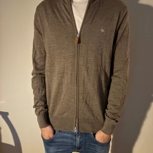 Brun kofta med dragkedja och brodyr - Säljer en brun kofta med dragkedja och diskret broderad logga på bröstet. Tröjan är långärmad och har ribbade muddar vid ärmslut och nederkant. Perfekt för lager-på-lager och chill vardagsstil. Storlek S