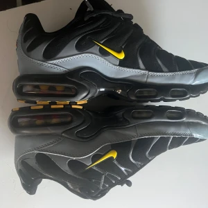 Nike Air Max Plus svart/grå med gul swoosh - Nike Air Max Plus sneakers i svart och grått med en snygg gul swoosh på sidan. Ovandelen är i mesh och syntet, med vågiga detaljer och synlig Air Max-dämpning i sulan. Yttersulan har gula inslag och mönstrad design för extra grepp. Perfekt för dig som gillar sportig och modern stil jag har använt som bara 2 gånger men säljer dom för dom är för små men. Jag köpte dom nya för 2599kr och säljer dom nu för 1600kr för att jag vill bara få solt dom annars står dom bara och dammar. Priset kan diskutera