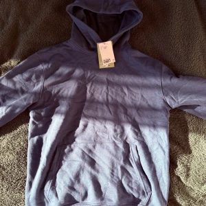 Blå hoodie med huva och magficka - Säljer en stilren blå hoodie från H&M med klassisk huva och stor magficka framtill. Perfekt för dig som gillar en enkel och avslappnad look. Tröjan har långa ärmar och är gjord i mjuk bomull för extra komfort.