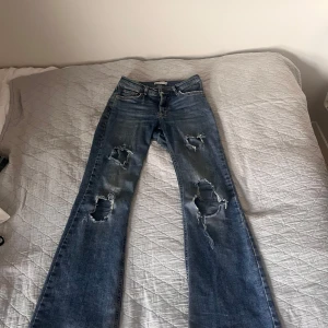 Blå bootcut jeans med slitningar - Bootcut jeans i klassisk blå tvätt från Gina tricot. Byxorna har stora slitningar och hål på båda benen för en edgy look. Femficksmodell med normal passform och snyggt utsvängda ben. Perfekta för dig som gillar en avslappnad och trendig stil.