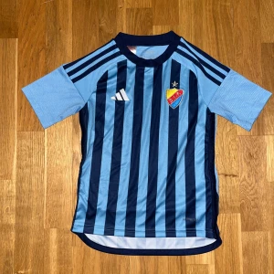Djurgården fotbollströja Adidas blå (128cm) - Djurgårdens IF fotbollströja från Adidas med klassiska ljus- och mörkblå vertikala ränder framtill, klubbmärke på bröstet och tre ränder på axlarna. Tillverkad i lätt och ventilerande syntetmaterial, perfekt för match eller träning.