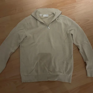 Beige stickad half zip från selected Homme - Beige stickad tröja från selected Homme med half zip-dragkedja i halsen. Tröjan har långärmad passform, ribbade muddar och en mjuk, strukturerad yta. Perfekt för lager-på-lager och chill höst- eller vinterstil.
