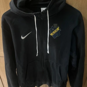 Svart AIK hoodie från Nike - Svart hoodie från Nike med AIK-logga på bröstet och vit snörning i huvan. Tröjan har dragkedja framtill, två fickor och klassisk Nike-swoosh. Perfekt för dig som vill visa ditt AIK-stöd med stil.
