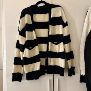 Randig svartvit kofta med knappar - Mysig kofta med breda svarta och vita ränder. Den har knappar framtill, två fickor och är stickad i ett mjukt material. 